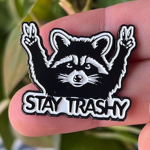Puede incluir: Pin de esmalte negro y blanco con un mapache con las patas levantadas en señal de paz. El mapache lleva una camisa a rayas blancas y negras y tiene una cara blanca con ojos negros. El texto "STAY TRASHY" está escrito en letras blancas debajo del mapache.