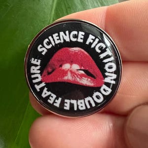 Op de afbeelding: Een ronde, zilveren pin met een zwarte achtergrond en witte tekst die "Double Feature Science Fiction" luidt. In het midden van de pin bevindt zich een rode, open mond met tanden.