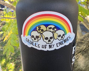 May include: A matte black water bottle featuring a sticker with a colorful rainbow arching over a pile of beige skulls. The text "SKULLS OF MY ENEMIES" is printed in black capital letters on a white cloud shape beneath the skulls.