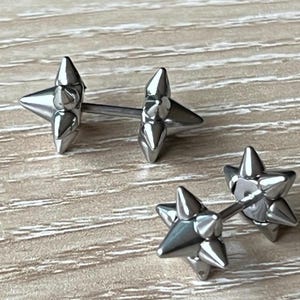 May include: Three pairs of silver-colored star-shaped stud earrings. Each earring features a star design with pointed edges. The earrings are displayed on a light brown wooden surface.