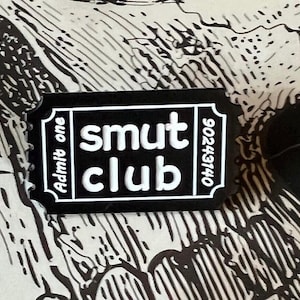 May include: Black and white ticket-shaped pin with the text "smut club" and "Admit one" on the left side and "902443140" on the right side.