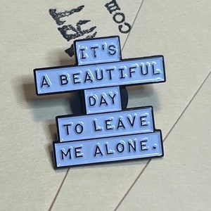 "it's a beautiful day to leave me alone." this is a soft enamel pin that is white with blacks words.