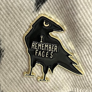 May include: A black enamel pin shaped like a crow with gold accents. The crow has the words "I Remember Faces" written on its body.