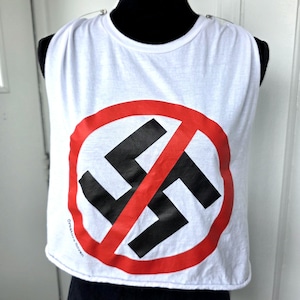 Op de afbeelding: Een witte mouwloze crop top met een grote grafische print op de voorkant. Het ontwerp bestaat uit een zwart hakenkruis in een rode cirkel met een rode diagonale streep erdoorheen. De tekst Creators Haven is in kleine zwarte letters gedrukt.