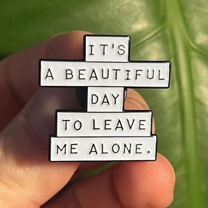 "it's a beautiful day to leave me alone." this is a soft enamel pin that is white with blacks words.