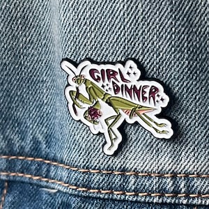 Pu&ograve; includere: Una mantide religiosa verde con occhi rossi tiene un cuore rosa nelle sue zampe. La mantide &egrave; su uno sfondo bianco con il testo "GIRL DINNER" in lettere rosa e nere.