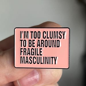 May include: A rectangular enamel pin with a pink background and black border. The pin features the text "I'M TOO CLUMSY TO BE AROUND FRAGILE MASCULINITY" in bold, black capital letters.