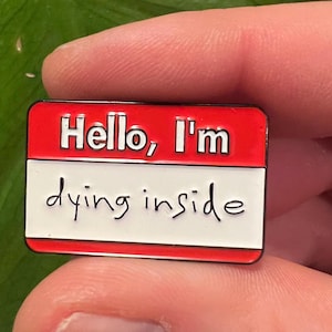 Peut inclure: Épinglette en émail rouge et blanc avec le texte "Hello, I'm dying inside" sur une étiquette de nom.