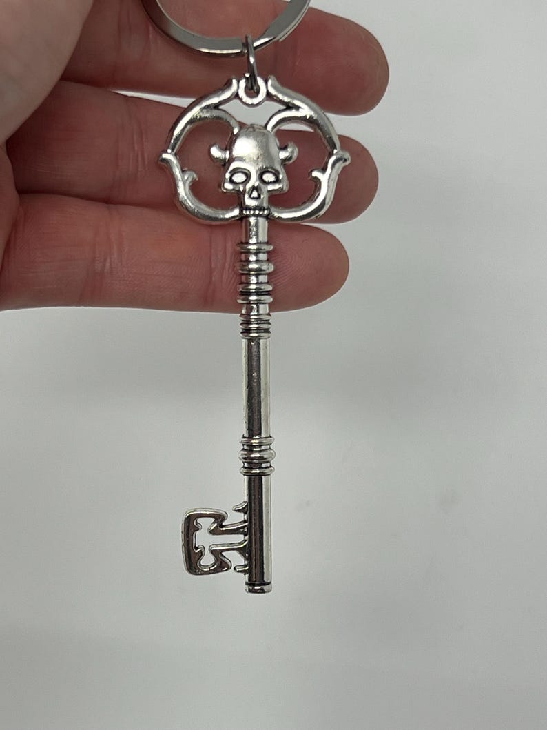 May include: Silver-colored key with a skull design on the head. The key has a decorative, ornate handle and a key ring at the top. The key's design is detailed, with a vintage or gothic aesthetic.