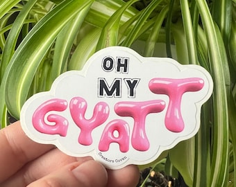 May include: Pink and white sticker with the text "Oh My GyaT" in a bubble font. The sticker is shaped like a cloud and has the text "Creators Coven" at the bottom.