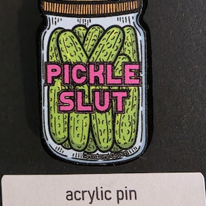 Könnte beinhalten: Acryl-Pin mit einem Glas Gurken mit dem Text "PICKLE SLUT" in Pink. Das Glas hat einen gelben Deckel und ist mit grünen Gurken gefüllt. Der Pin befindet sich auf einer schwarzen Oberfläche. Der Text "acrylic pin" steht darunter.