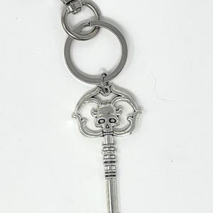 May include: Silver-colored key chain featuring a decorative key pendant with a skull design. The key has a detailed, ornate pattern and is attached to a key ring and a clasp. The key chain is made of metal and is suitable for keys.