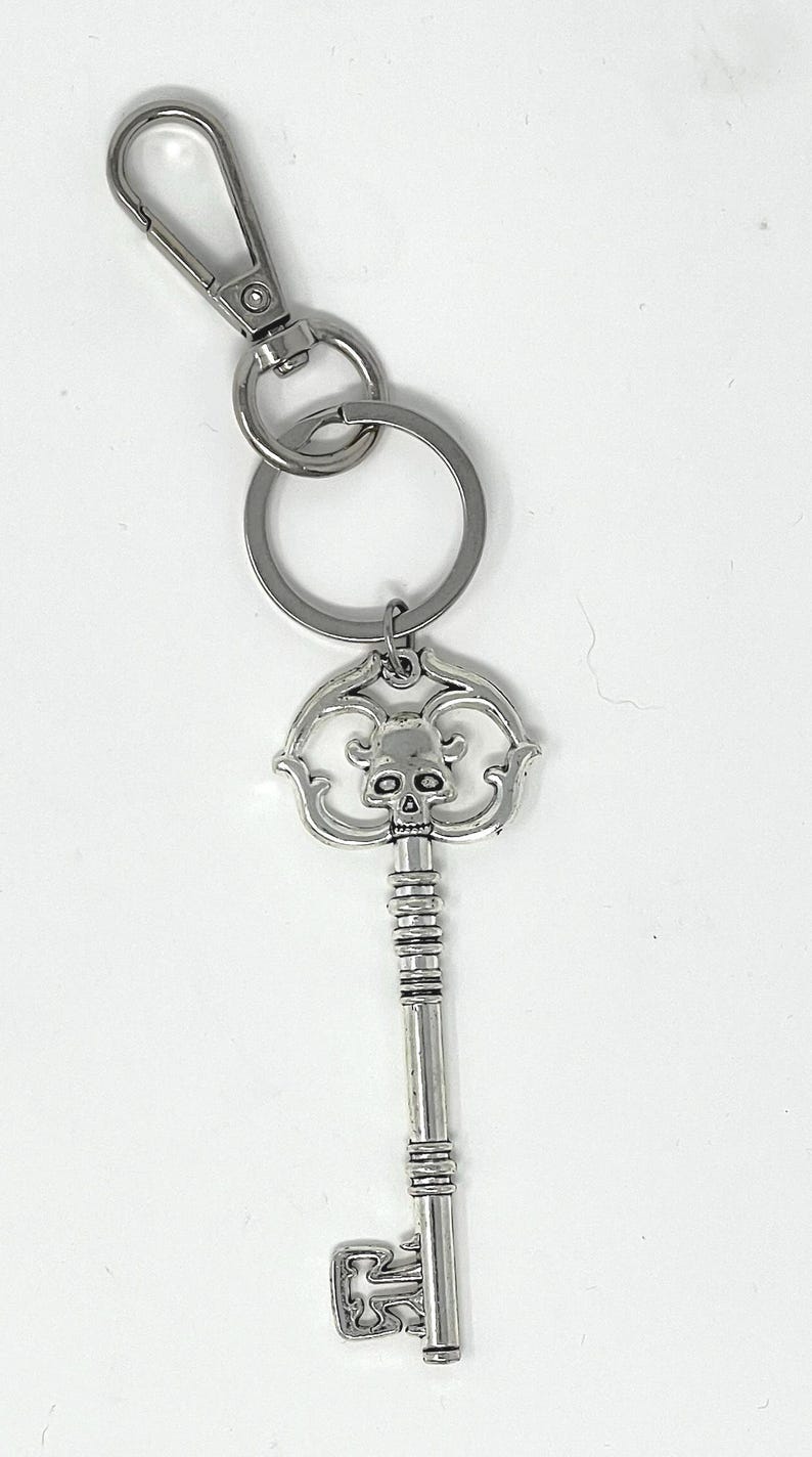 May include: Silver-colored key chain featuring a decorative key pendant with a skull design. The key has a detailed, ornate pattern and is attached to a key ring and a clasp. The key chain is made of metal and is suitable for keys.