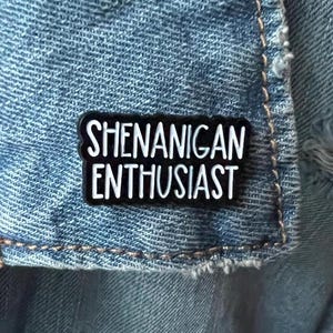 May include: Black enamel pin with white text that reads "Shenanigan Enthusiast".