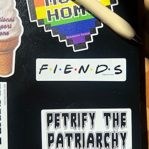 May include: Three stickers on a black surface. The first sticker is a rainbow heart with the text "Move Home" and "Creators Coven". The second sticker is white with black text that reads "F.I.E.N.D.S" and "Creators Coven". The third sticker is white with black text that reads "Petrify the Patriarchy" and "Creators Coven".