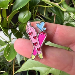 May include: A pink and green enamel pin featuring two crossed joints with hearts on them. The joints are topped with a blue heart.