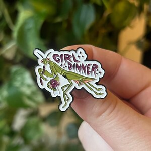 Pu&ograve; includere: Uno spillo in smalto bianco con una mantide religiosa verde che tiene un insetto viola pi&ugrave; piccolo. Lo spillo dice "Girl Dinner" in un carattere viola con stelle.
