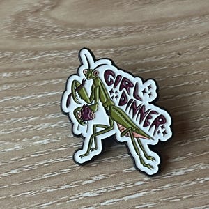 Pu&ograve; includere: Una mantide religiosa verde con le gambe rosa che tiene un oggetto viola nelle sue zampe anteriori. La mantide &egrave; su uno sfondo bianco con il testo "Girl Dinner" in lettere marroni.