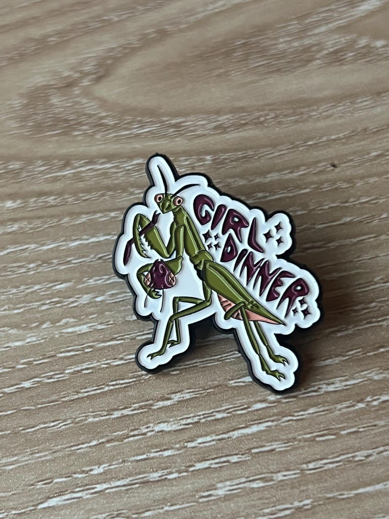 Pu&ograve; includere: Una mantide religiosa verde con le gambe rosa che tiene un oggetto viola nelle sue zampe anteriori. La mantide &egrave; su uno sfondo bianco con il testo "Girl Dinner" in lettere marroni.
