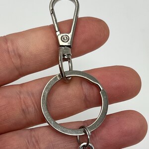 May include: Close-up of a silver-colored keychain with a lobster clasp, a split ring, and a decorative charm. The keychain is made of metal and is designed to hold keys or other small items. The background is a light color.