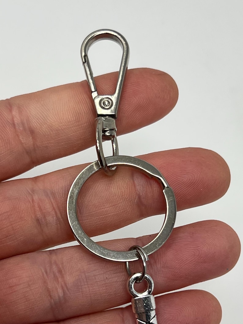 May include: Close-up of a silver-colored keychain with a lobster clasp, a split ring, and a decorative charm. The keychain is made of metal and is designed to hold keys or other small items. The background is a light color.