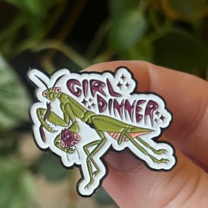 Könnte beinhalten: Eine weiße Emaille-Anstecknadel mit einer grünen Gottesanbeterin mit rosa Augen. Die Gottesanbeterin hält ein kleineres, violettes Insekt in ihren Vorderbeinen. Der Text "GIRL DINNER" ist in lila Buchstaben über der Gottesanbeterin geschrieben.