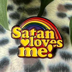May include: A rainbow enamel pin with the text "Satan loves me!" in yellow letters with a red heart in the middle of the word "loves".