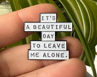 May include: White enamel pin with the text "It's a beautiful day to leave me alone."