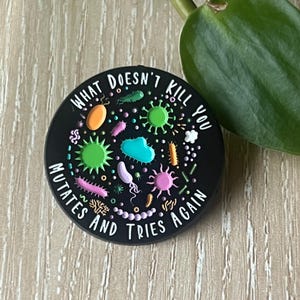 Puede incluir: Soporte redondo negro para teléfono con ilustraciones de microbios coloridos y el texto "WHAT DOESN'T KILL YOU MUTATES AND TRIES AGAIN". El diseño presenta varias formas y colores, incluyendo verde, rosa y azul.