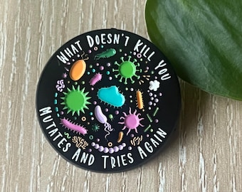 Può includere: Supporto rotondo nero per telefono con illustrazioni di microbi colorati e la scritta "WHAT DOESN'T KILL YOU MUTATES AND TRIES AGAIN". Il design presenta varie forme e colori, tra cui verde, rosa e blu.