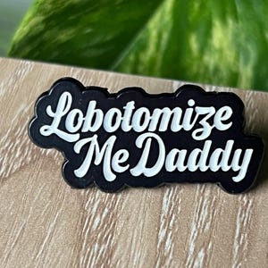May include: Black enamel pin with white lettering that reads "Lobotomoize Me Daddy".