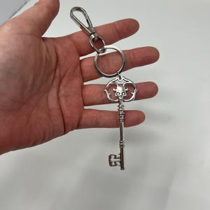 May include: Silver-colored key chain with a decorative key pendant. The key features a skull design at the top and a detailed, ornate shaft. Includes a metal clasp and a key ring for attaching to keys or a bag.