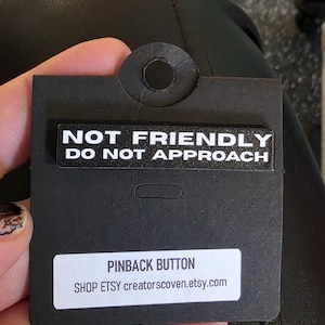 May include: A black pin badge with the white text "NOT FRIENDLY DO NOT APPROACH". The badge is on a black card with a white label that reads "PINBACK BUTTON".