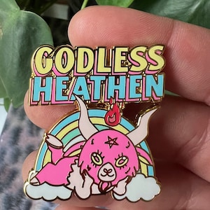 May include: A gold-coloured enamel pin with the words "GODLESS HEATHEN" in a colourful font. The pin features a pink cartoon goat with horns, a rainbow, and a flame. The pin is set against a blurred background of green leaves.