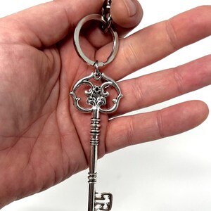 May include: Silver-colored key-shaped keychain. The key has an ornate design on the head and a long, detailed stem. A small ring and clasp are attached for securing keys. The keychain is held against a white background.