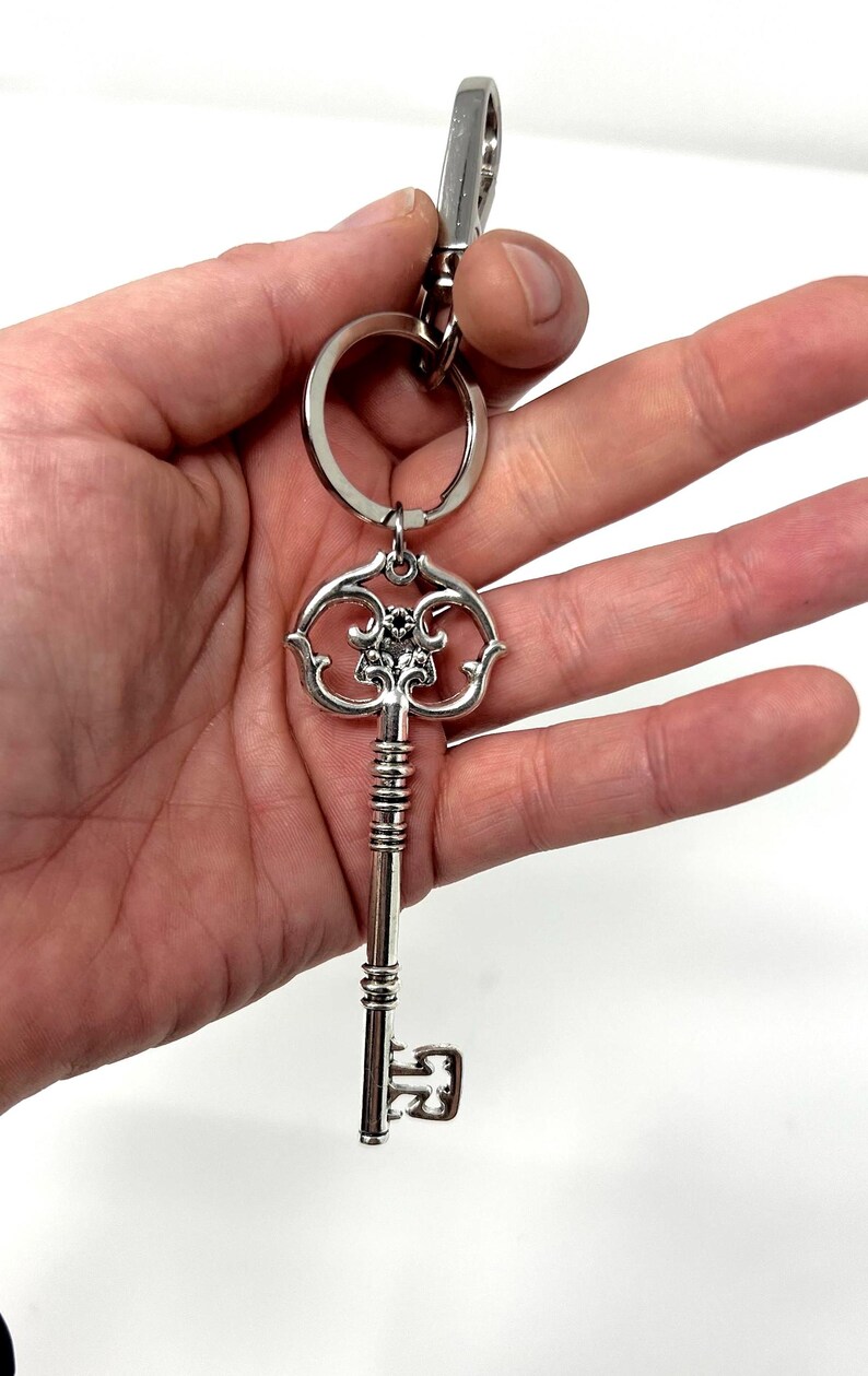 May include: Silver-colored key-shaped keychain. The key has an ornate design on the head and a long, detailed stem. A small ring and clasp are attached for securing keys. The keychain is held against a white background.