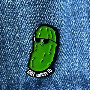 May include: A green enamel pin featuring a cartoon pickle wearing sunglasses. The pin has a black outline and the words "Dill with it" in white text. The pin is set against a blue denim background.