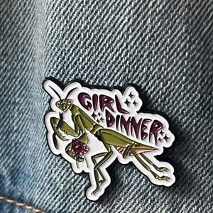 Pu&ograve; includere: Una mantide religiosa verde con le gambe rosa tiene una creatura viola pi&ugrave; piccola tra le braccia. La mantide &egrave; su uno sfondo bianco con il testo "Girl Dinner" in lettere nere e viola.