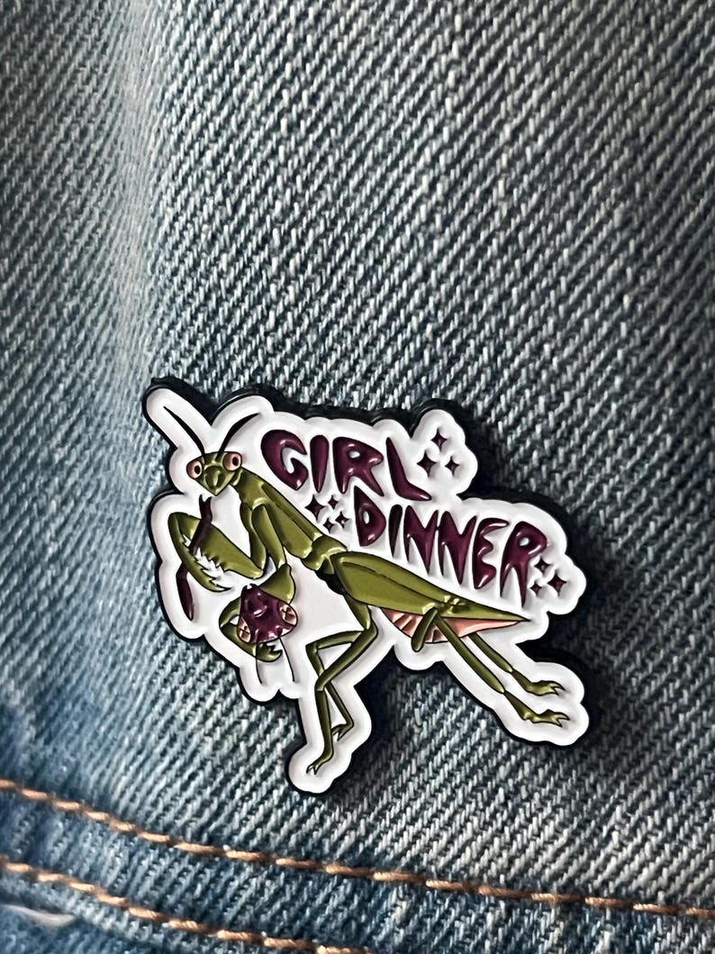 Pu&ograve; includere: Una mantide religiosa verde con le gambe rosa tiene una creatura viola pi&ugrave; piccola tra le braccia. La mantide &egrave; su uno sfondo bianco con il testo "Girl Dinner" in lettere nere e viola.