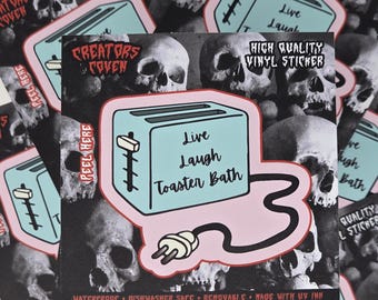 May include: A square vinyl sticker featuring a light blue toaster with a black power cord and plug. The toaster has the text "Live Laugh Toaster Bath" printed on its side. The background consists of a black and white pattern of human skulls.