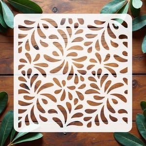 May include: A square white stencil featuring a repeating pattern of stylized, curved leaf-like shapes arranged in a flowing, organic design. The stencil has rounded corners and cutouts that create a symmetrical, botanical-inspired motif.