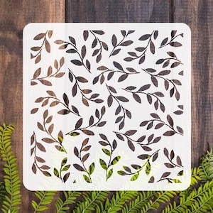 May include: A white square stencil featuring a repeating pattern of stylized leaves and branches in shades of brown and black. The stencil is placed on a dark wood surface, with green foliage visible at the bottom and sides.