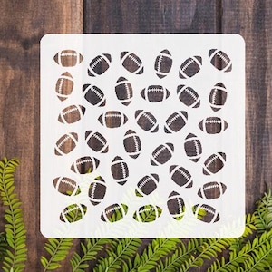 May include: A white stencil featuring a repeating pattern of footballs. The footballs are brown with white laces and are arranged in a scattered design. The stencil is on a dark wood background with green foliage.