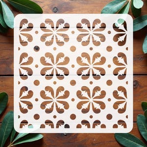 May include: A square stencil featuring a repeating pattern of four-petaled floral motifs and small circular dots. The design is cut out from a flat, off-white material, creating a symmetrical grid of decorative shapes across the entire surface.