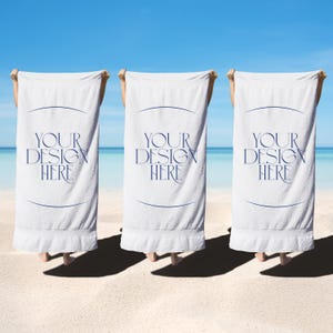 May include: Three white beach towels with the text "YOUR DESIGN HERE" in navy blue. The towels are held up against a backdrop of a blue sky and ocean. The towels are on a sandy beach.