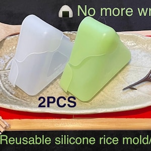 May include: Two silicone rice molds, one white and one green, on a plate with a wooden spoon and a small flower sprig. The text "No more wrap!" and "2PCS Reusable silicone rice mold/case" is visible in the image.