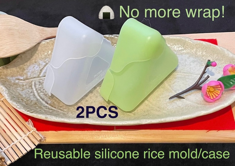 May include: Two silicone rice molds, one white and one green, on a plate with a wooden spoon and a small flower sprig. The text "No more wrap!" and "2PCS Reusable silicone rice mold/case" is visible in the image.
