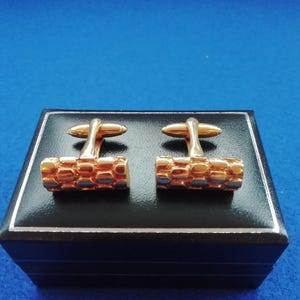 May include: A pair of gold cufflinks in a black box. The cufflinks have a textured, geometric design.