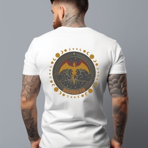 May include: White t-shirt with a circular graphic on the back featuring a winged creature in gold and red, surrounded by a ring of moons and stars. The word "BASGIATH" is printed below the creature in gold.