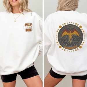May include: White sweatshirt with a front graphic that reads "FLY OR DIE" in a brown and gold design. The back features a circular design with a phoenix, clouds, and the word "BASUATH".